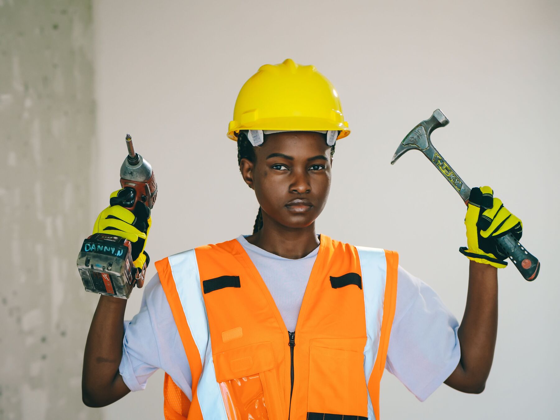 A site worker in a yellow hard hat and orange vest holding a drill and a hammer